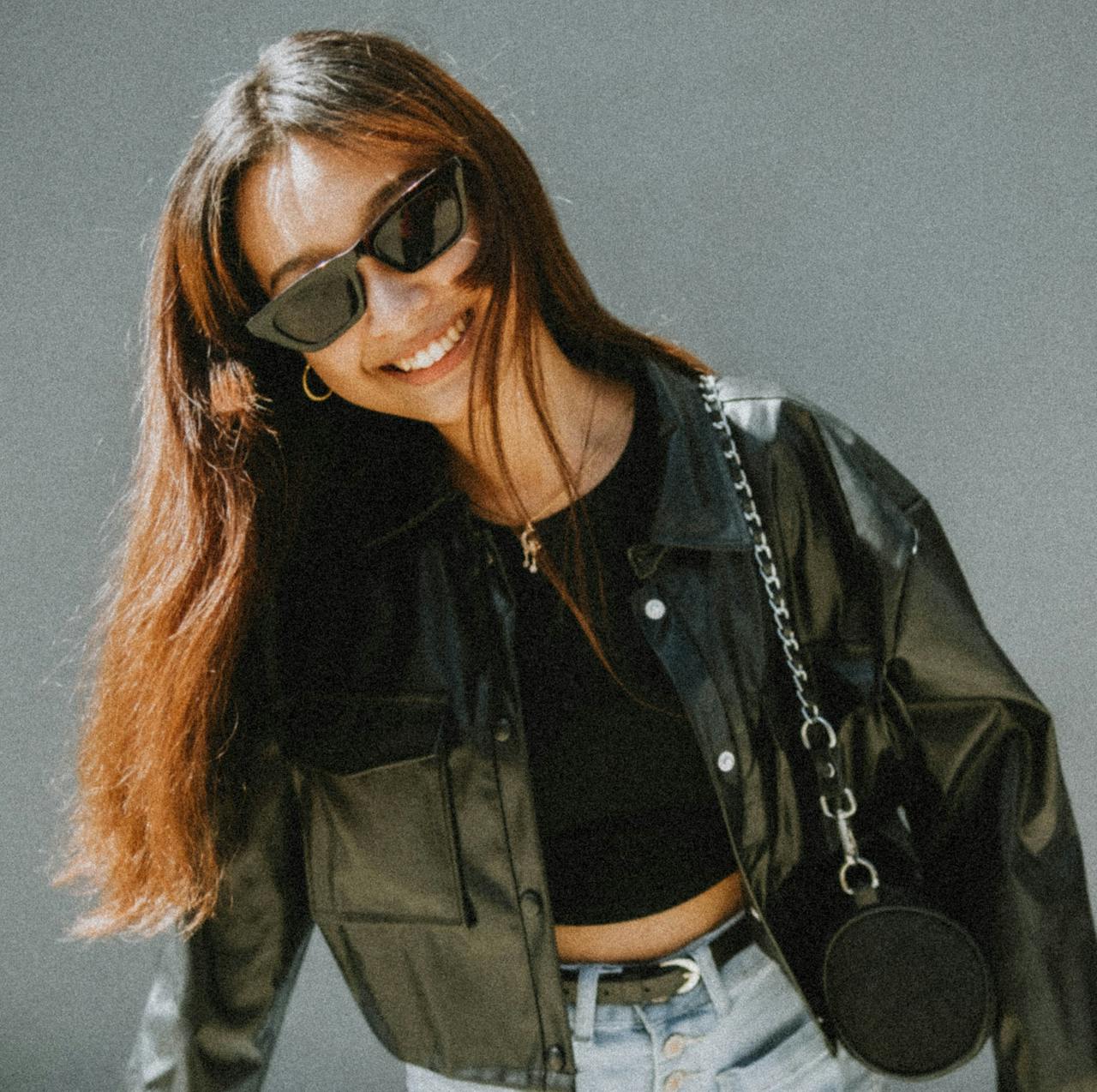 Stylish woman wearing sunglasses and a leather jacket, smiling confidently outdoors.