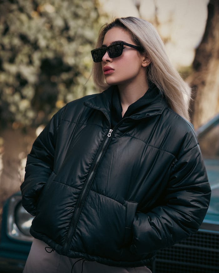 Blonde woman wearing black puffer jacket and sunglasses, posing outdoors with a confident look.