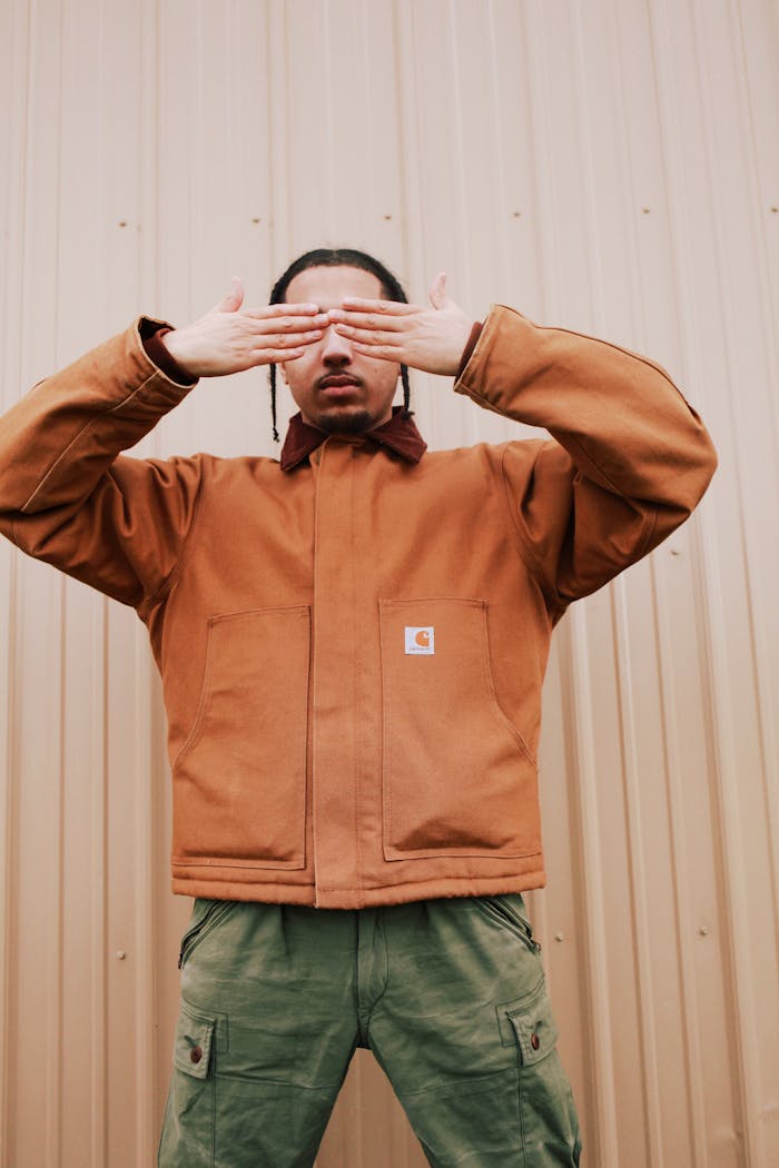 A man covers his face with his hands while posing in a stylish outfit against a beige wall.